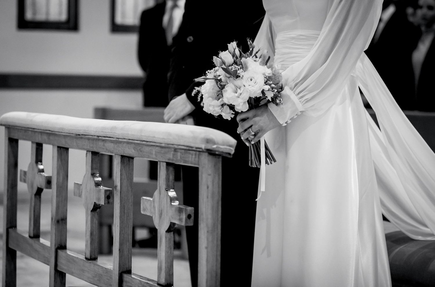 Black and white photo of a bride holding a bouquet standing beside wooden church pews during a wedding ceremony.