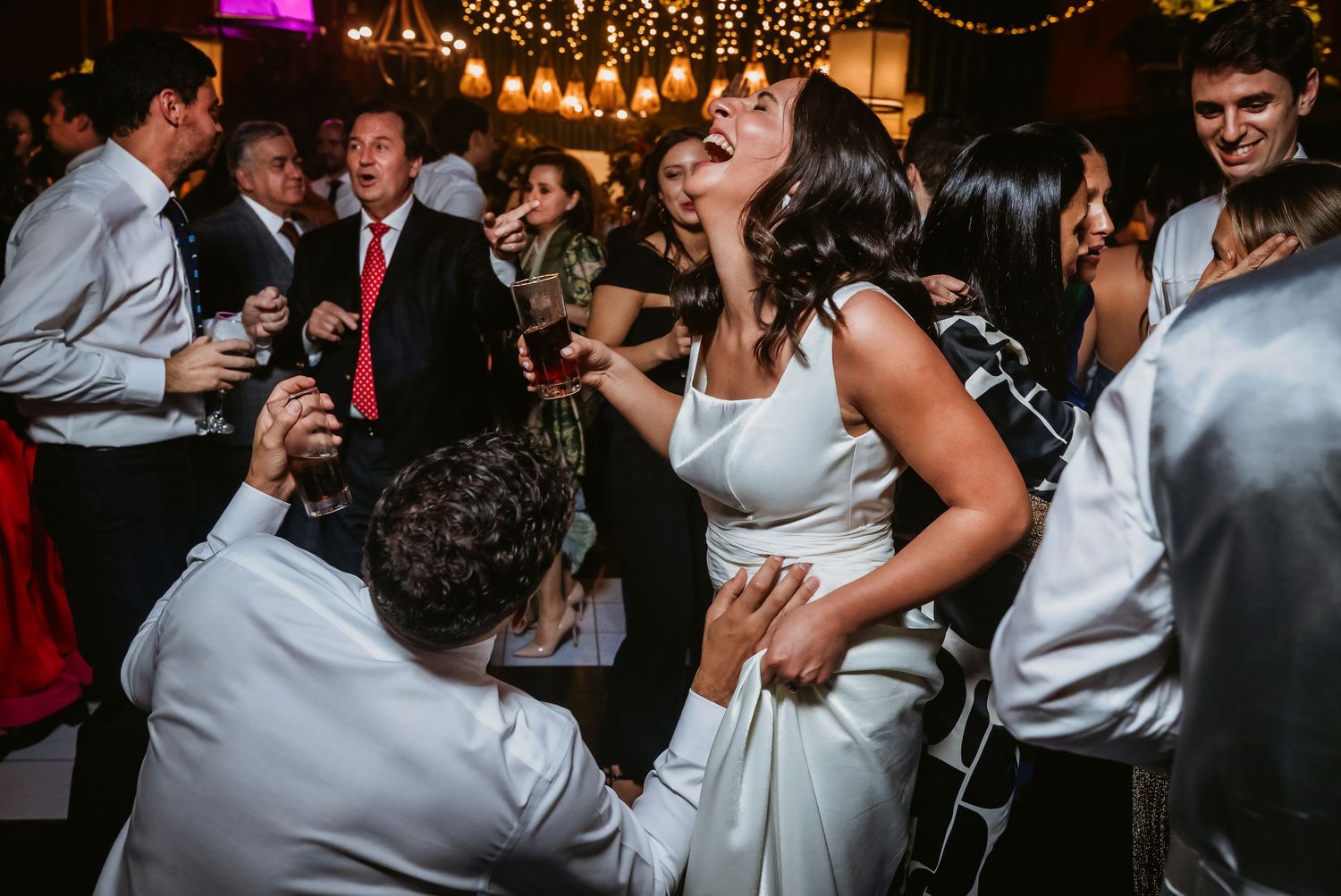Bride laughing and dancing joyfully with guests at a lively wedding reception under warm string lights.