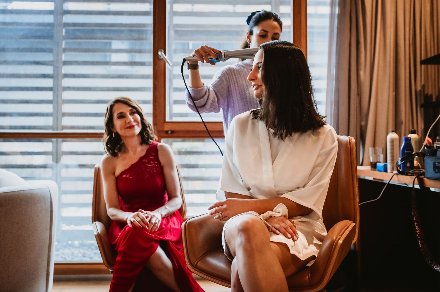 Woman in red dress watches hairstylist blow-dry another woman's hair in a bright, elegant room.