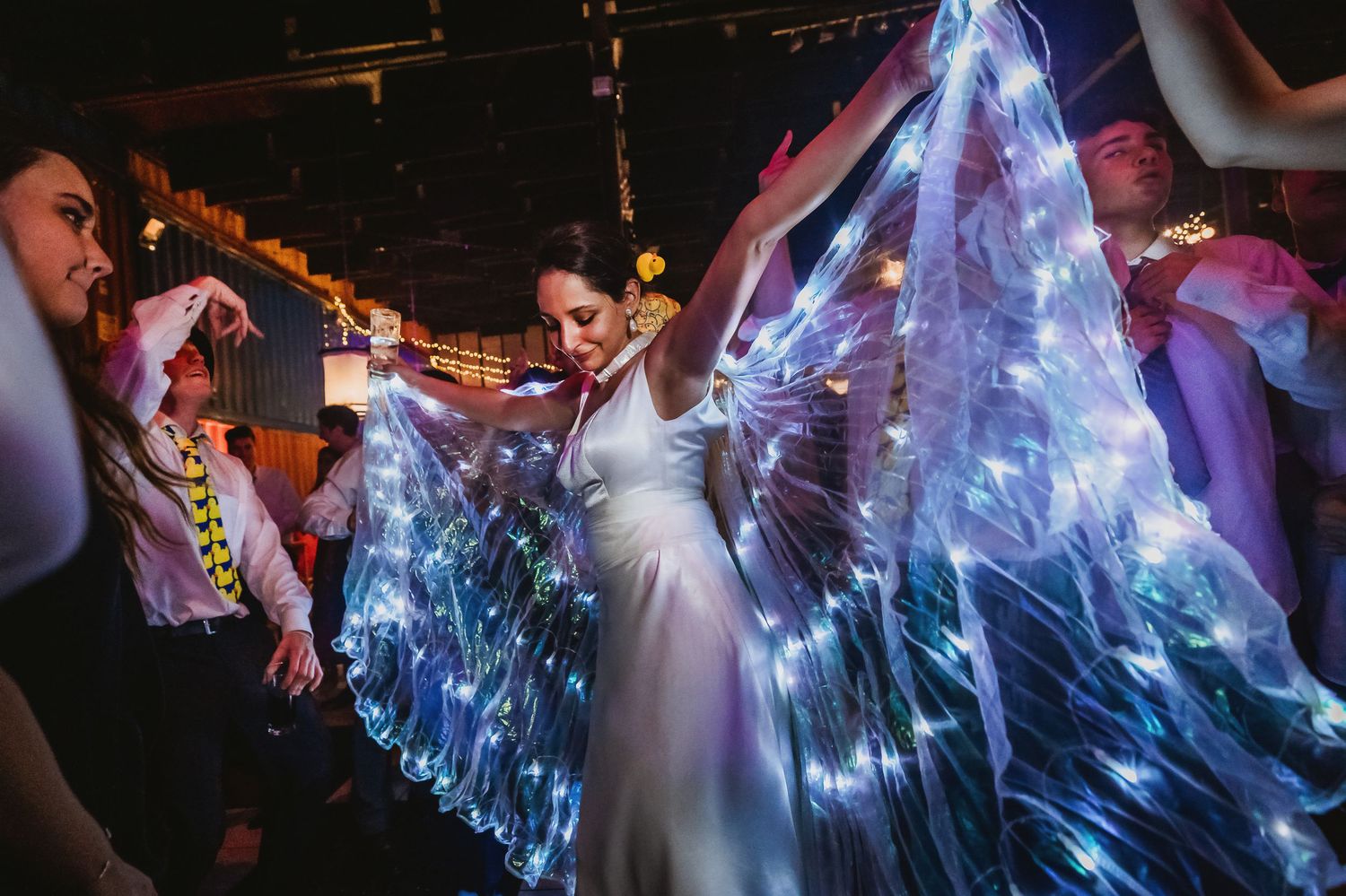 Bride dancing in white dress wearing illuminated LED light-up butterfly wings at a wedding reception party.