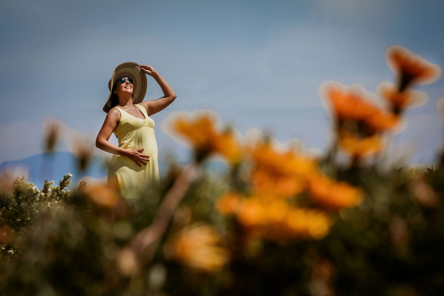 Pregnant woman in yellow dress and hat standing in a field of orange wildflowers under a blue sky.
