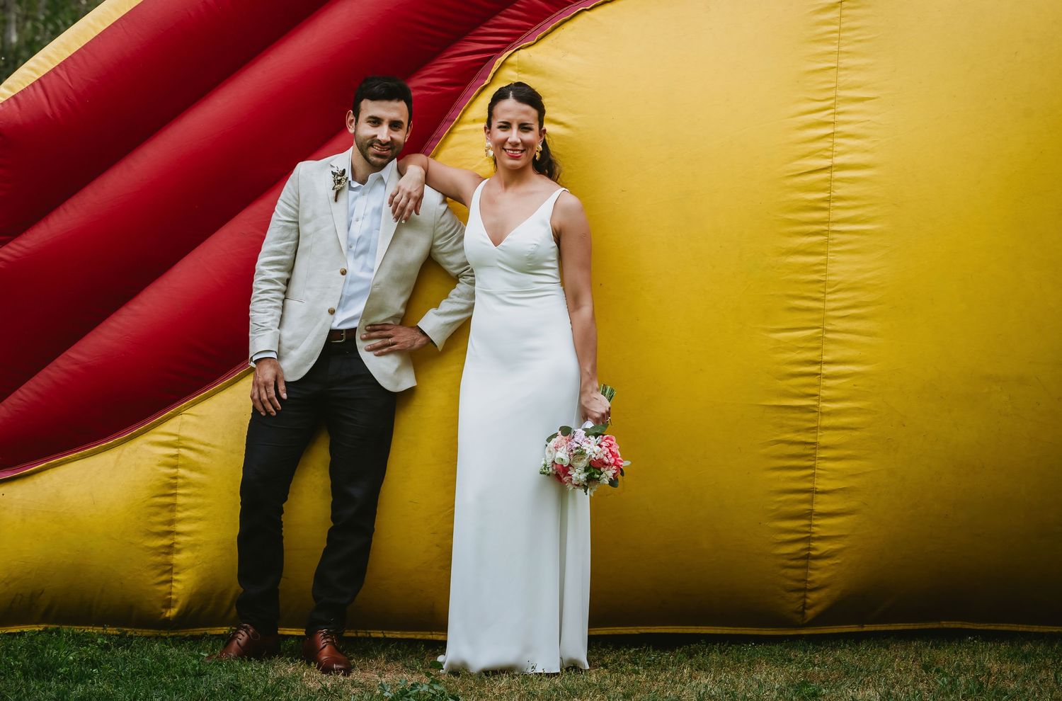 Bride and groom posing together in front of a colorful inflatable bounce house at their outdoor wedding.