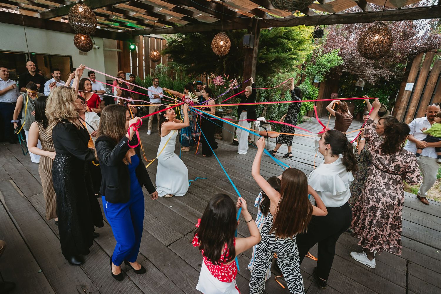 Wedding guests playing ribbon game on outdoor deck, bride in white dress surrounded by celebrating family and friends.