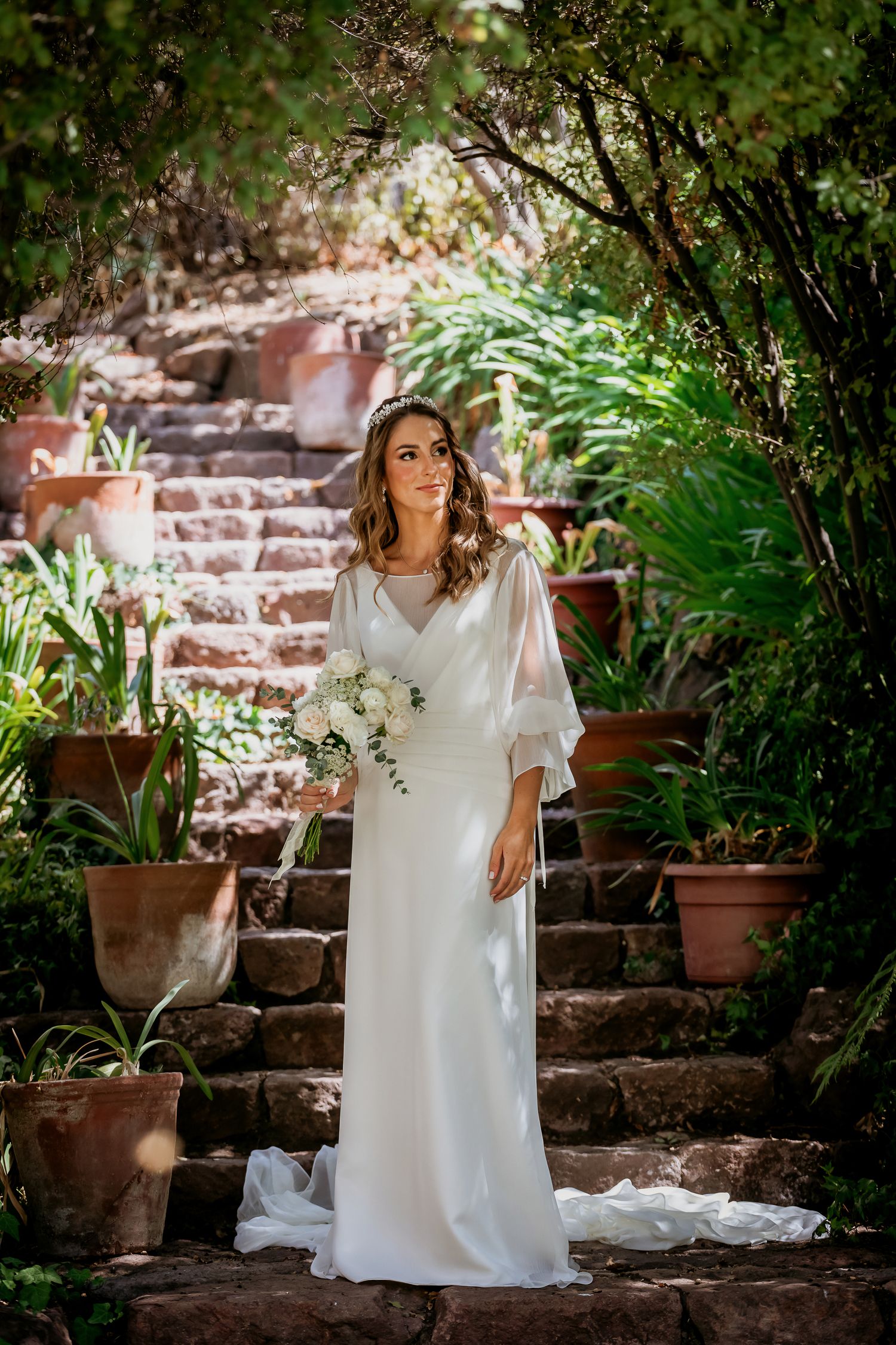Elegant bride in a flowing white gown holds a white bouquet on stone steps in a lush garden.