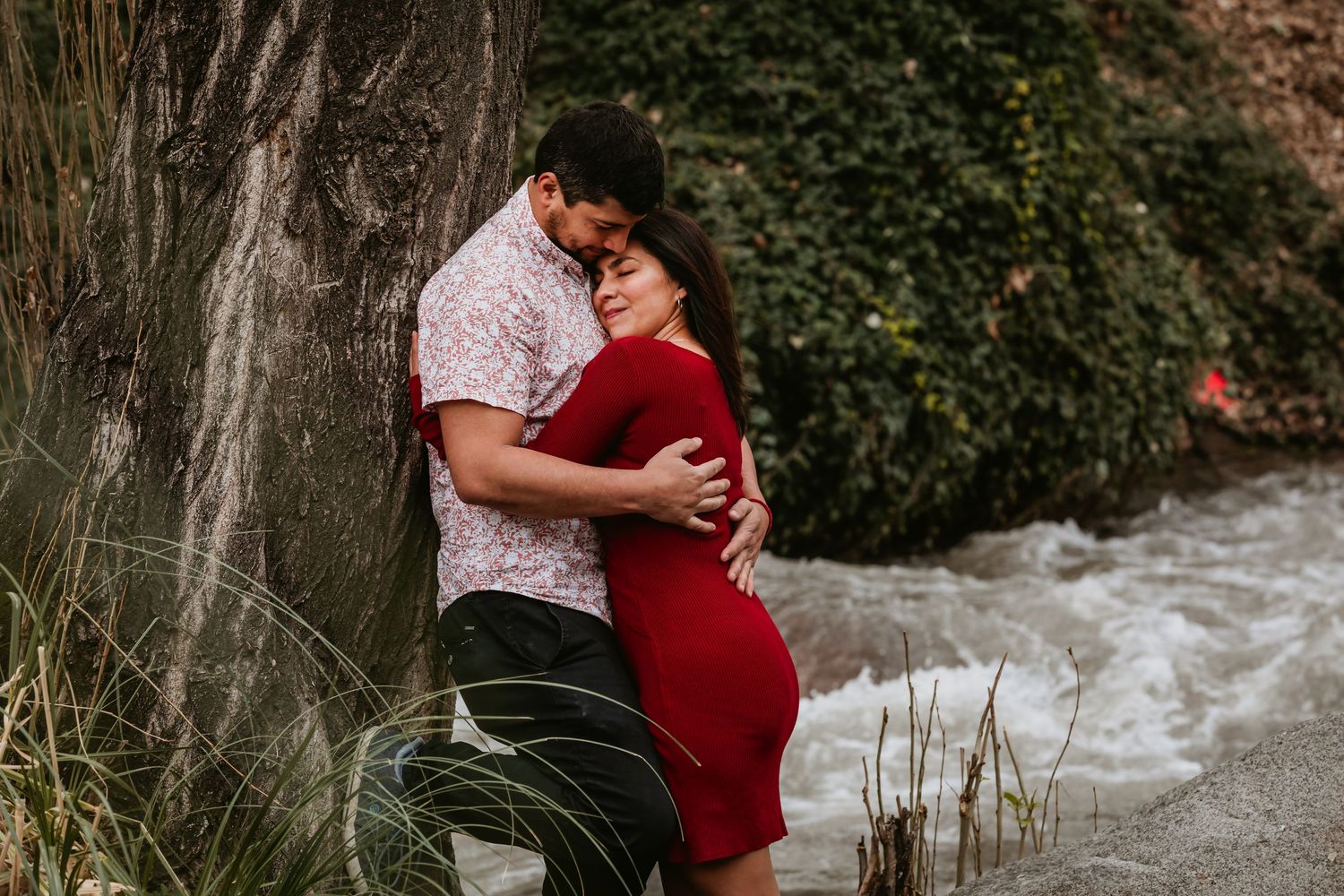 Couple embracing romantically against a tree near a rushing stream, woman in red dress.
