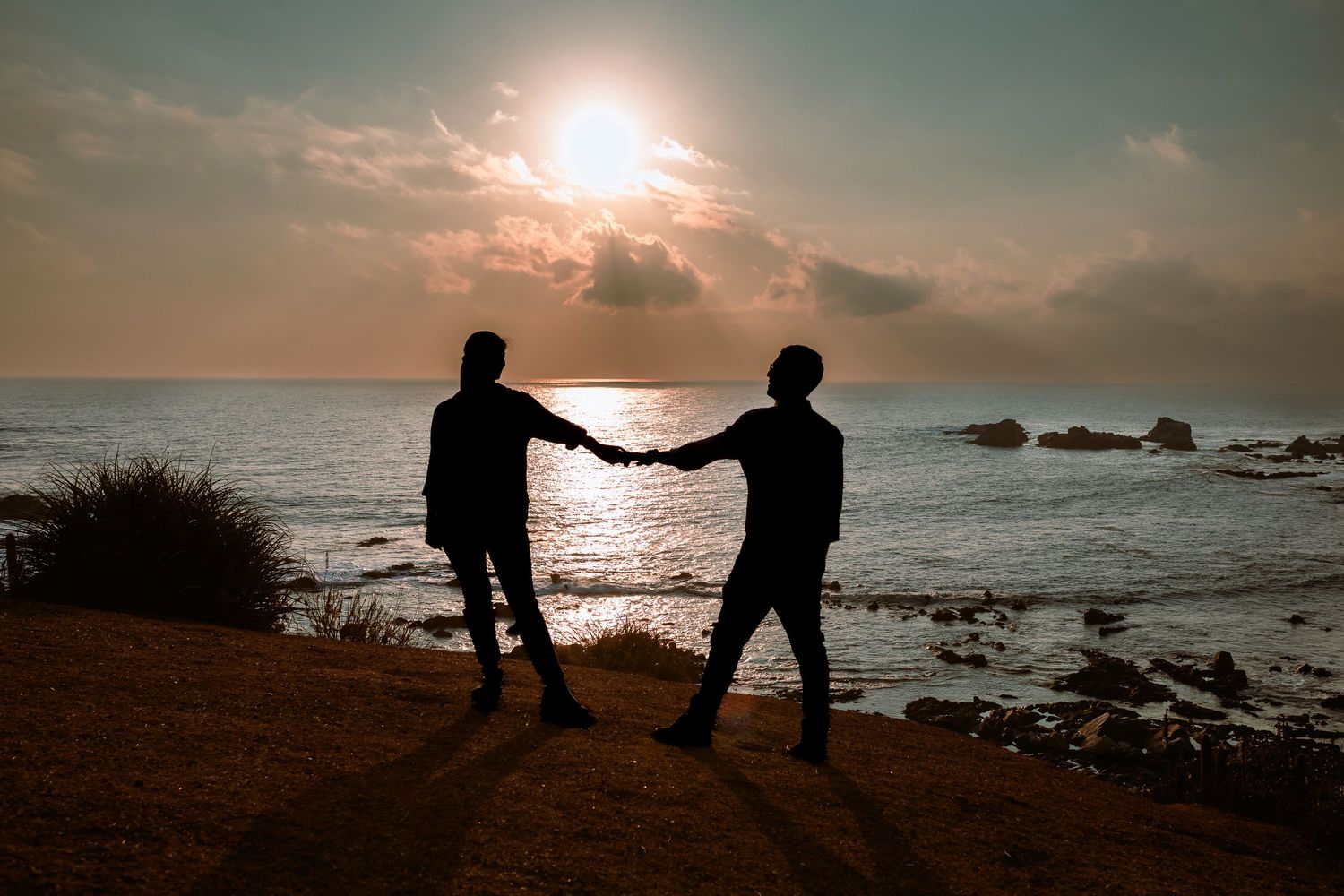 Silhouette of couple holding hands against glowing sunset over rocky ocean shore.