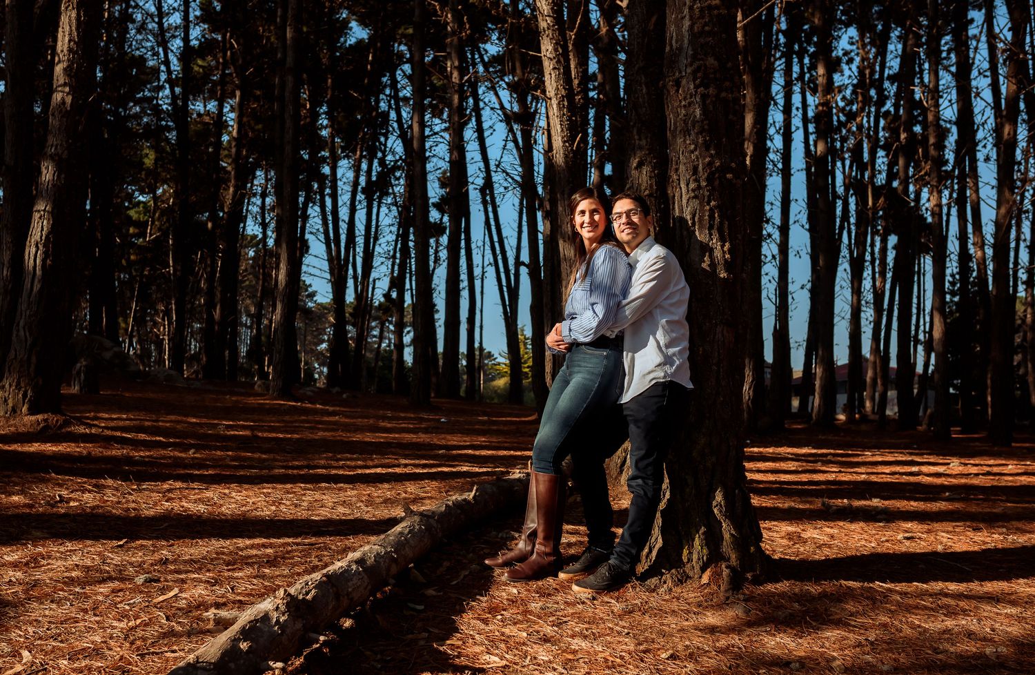 Couple leaning against pine tree trunk in sunlit forest with pine needle covered ground during engagement photo session.