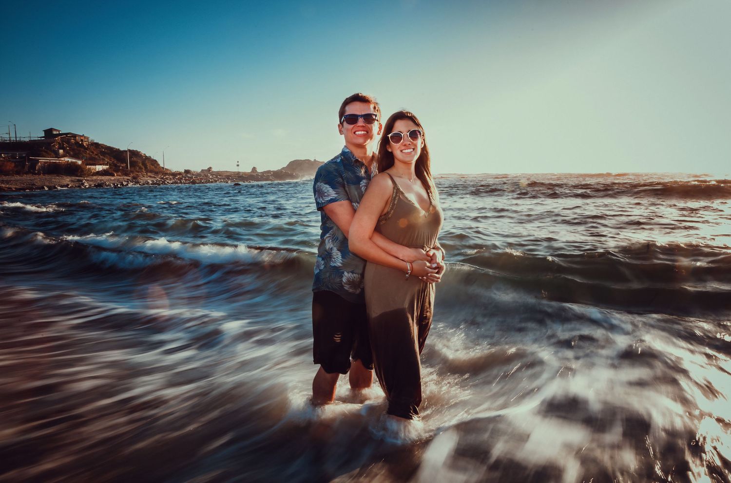 Couple hugging in ocean waves with a rocky coastal hillside in the background.