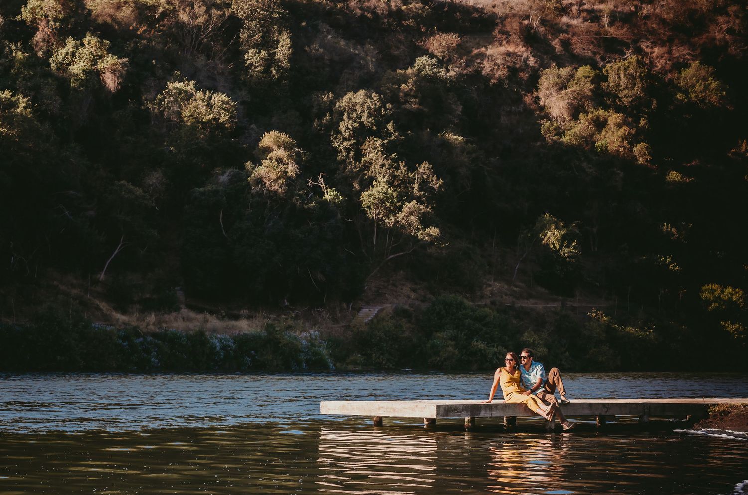 Couple sitting together on a floating dock surrounded by a calm lake and forested hills.