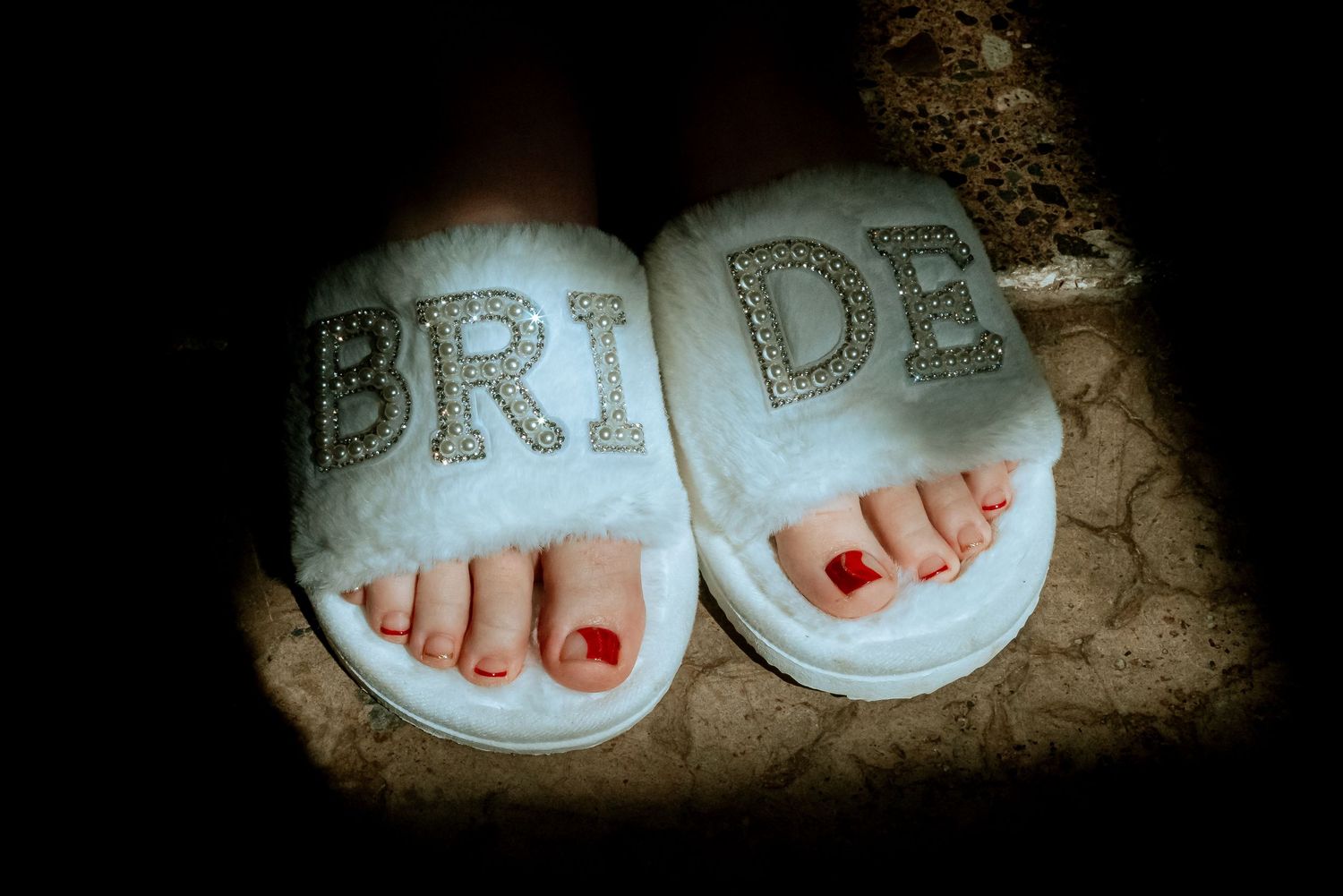 Close-up of bride's feet wearing white fluffy BRIDE rhinestone slippers with red pedicure.