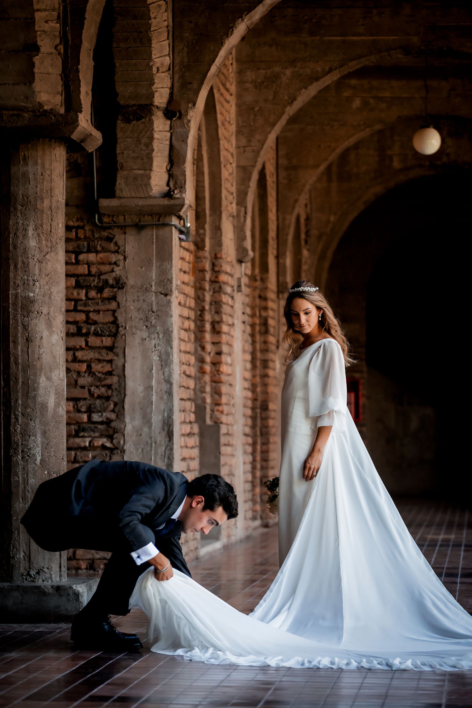 Groom adjusting bride's elegant white gown train in a dramatic brick archway setting.