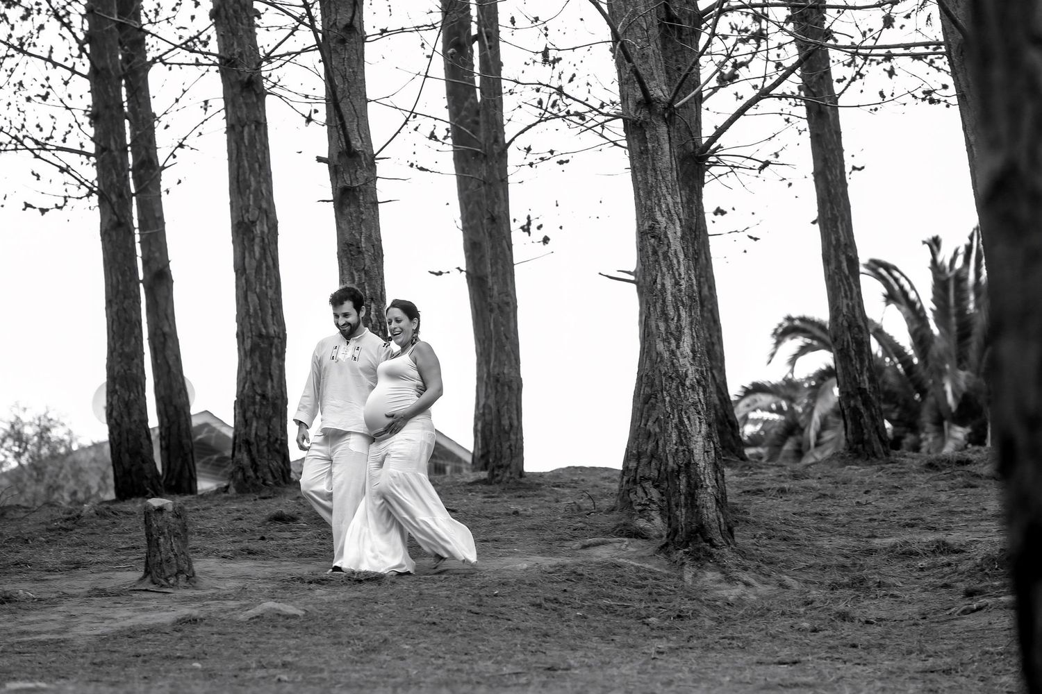 Black and white photo of a couple walking among tall trees, woman in white wedding dress, man in white shirt.