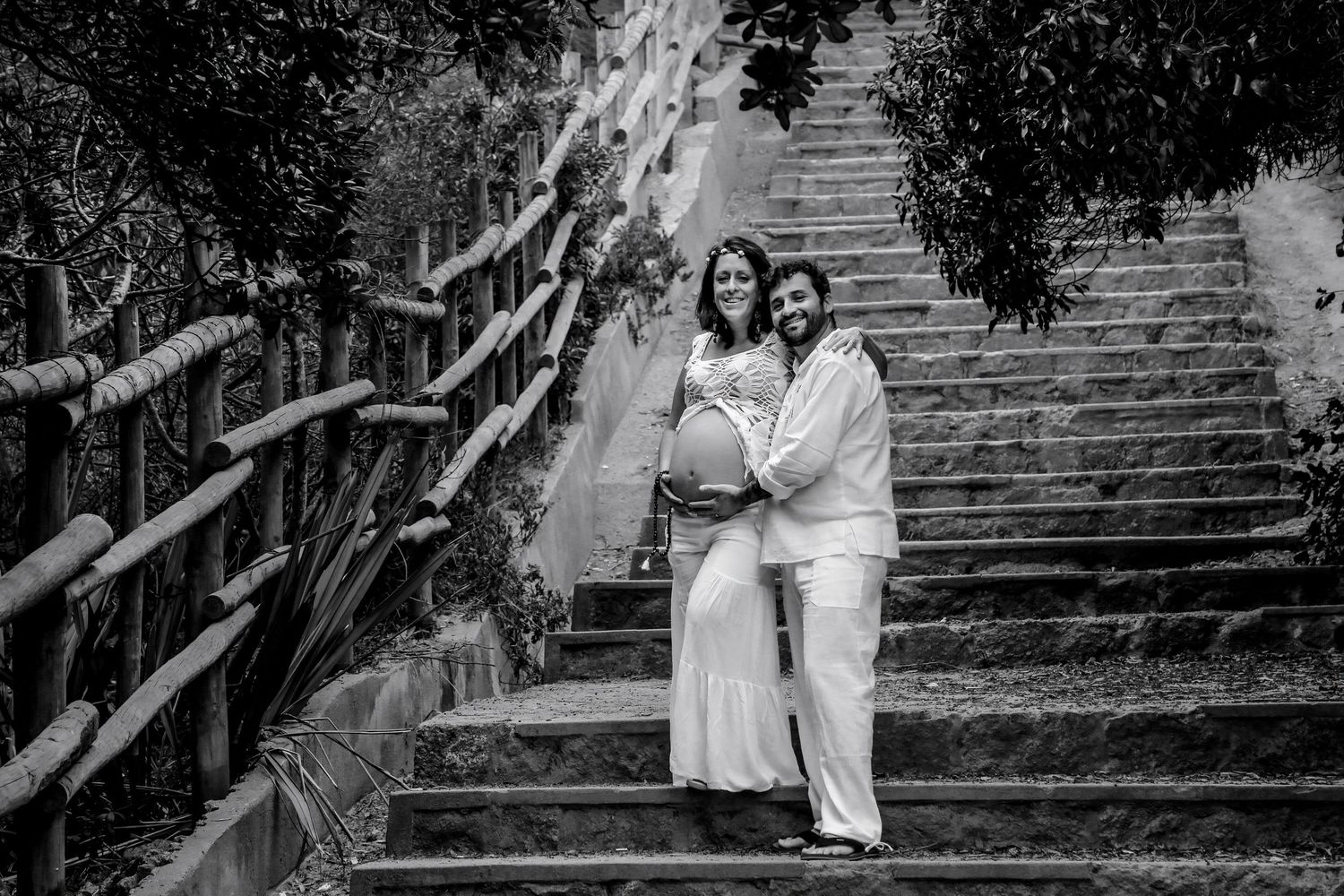 Black and white photo of a pregnant couple posing on outdoor stone steps surrounded by lush trees.