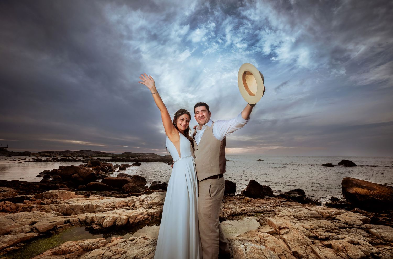 Joyful couple celebrating on rocky ocean shore, woman in blue gown and man in suit raising arms under dramatic cloudy sky.