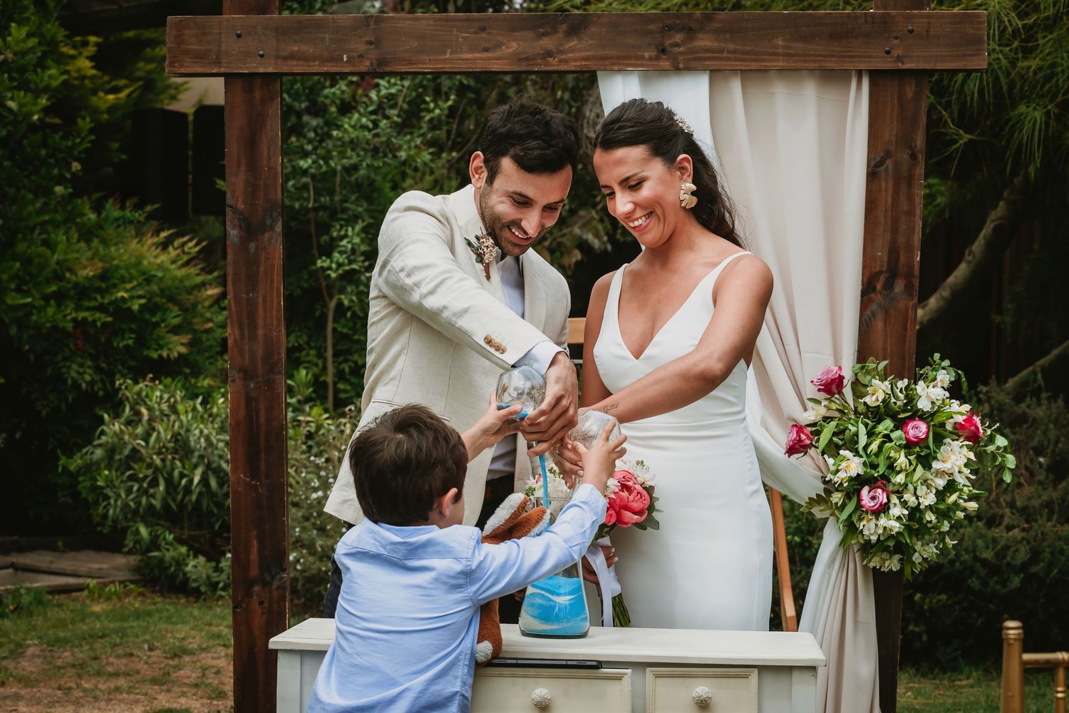 Bride and groom exchange rings with child at outdoor wedding ceremony under wooden arch with floral decorations.