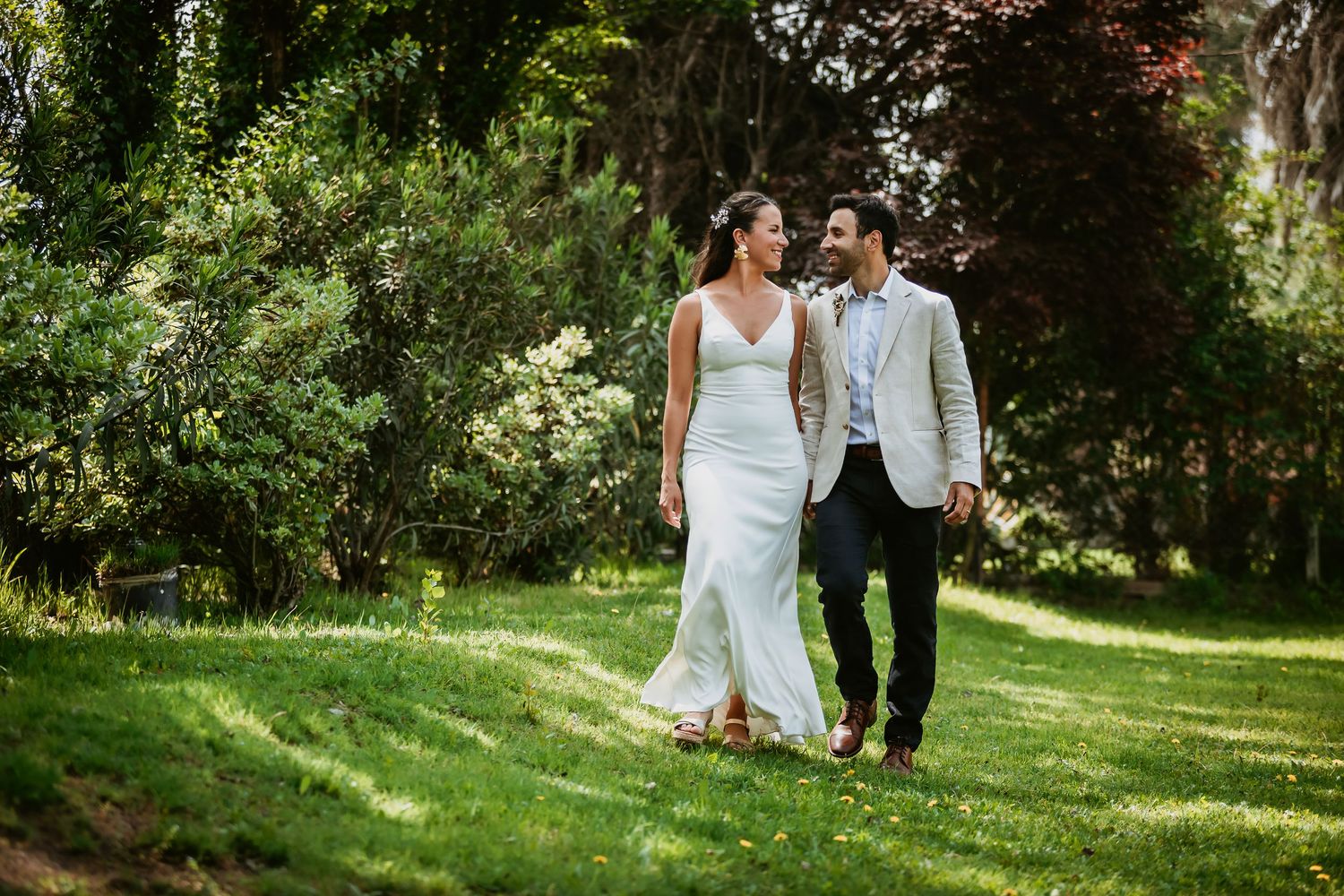 A bride and groom walk hand in hand through a lush green garden on their wedding day.