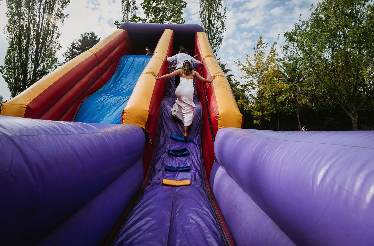 Child sliding down a colorful inflatable bounce house slide outdoors surrounded by green trees.
