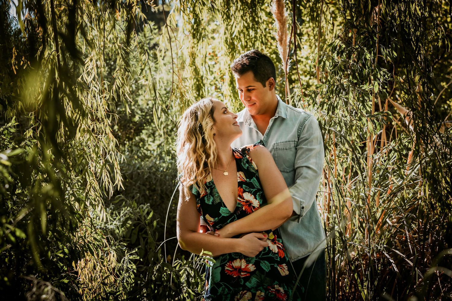 Couple embracing romantically among lush green weeping willow trees in a sun-dappled outdoor engagement photo session.
