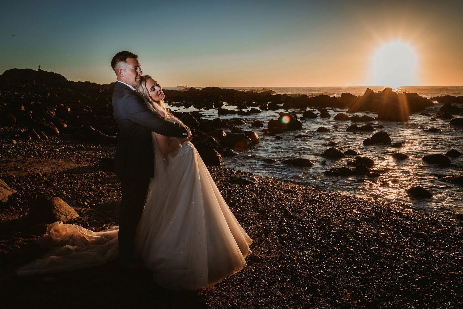 Couple embracing romantically on rocky shore with waves and glowing sunset behind them.