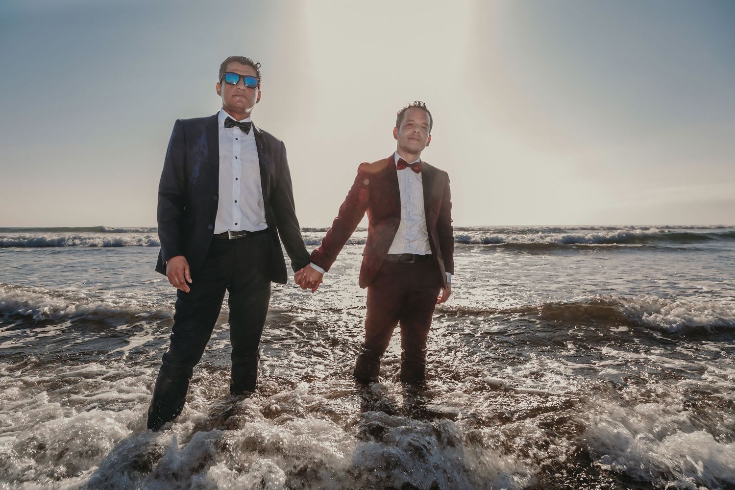 Two grooms in tuxedos hold hands while standing in ocean surf at sunset on their wedding day.