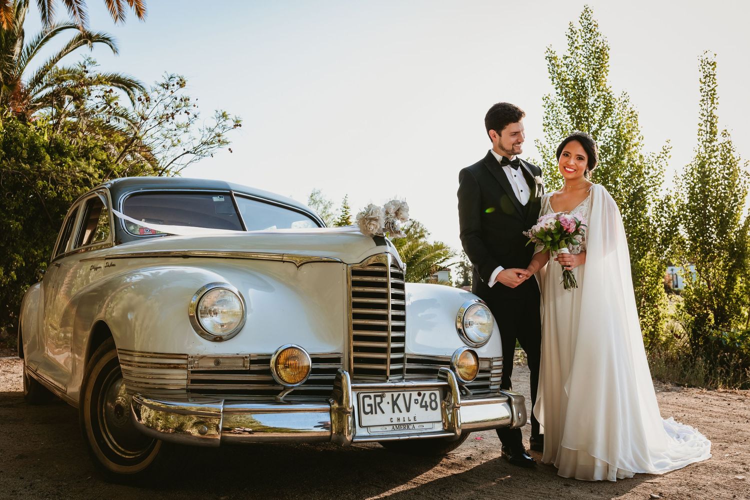 Bride and groom stand proudly beside decorated vintage white Packard classic car on sunny wedding day.