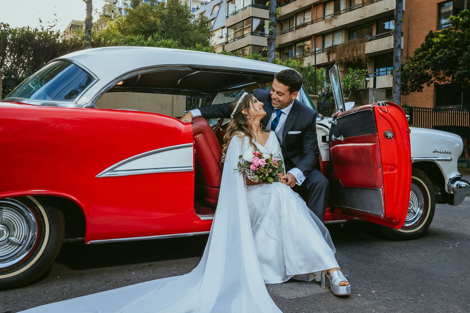 Newlyweds sharing a romantic moment leaning against a red vintage classic car with bridal bouquet.