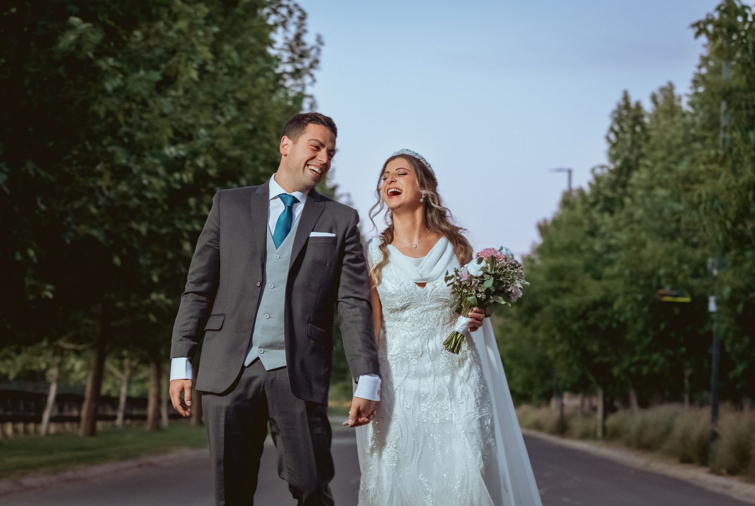 Happy bride and groom walk hand in hand laughing along a tree-lined pathway on their wedding day.