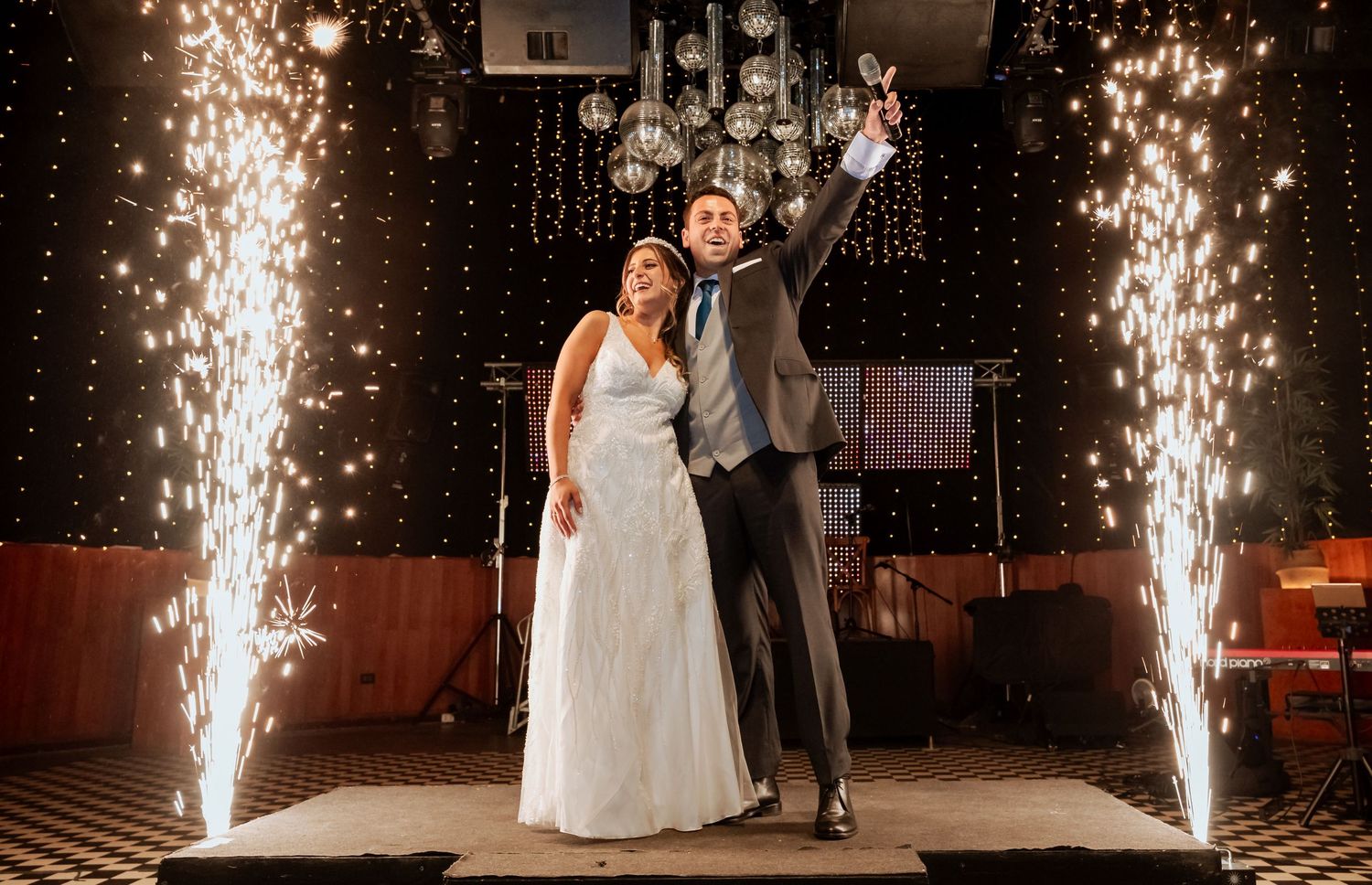 Newlyweds pose together on stage surrounded by sparkling fountain fireworks at their wedding reception.