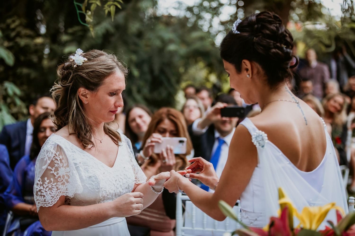 Two brides exchange wedding rings during an emotional outdoor garden ceremony.