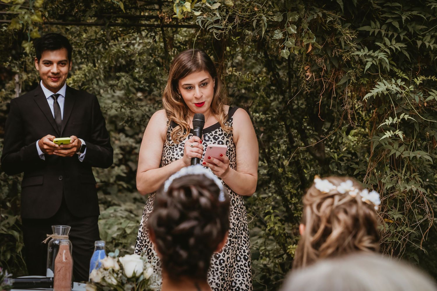 Woman in leopard print dress gives a heartfelt speech at an outdoor wedding reception surrounded by greenery.