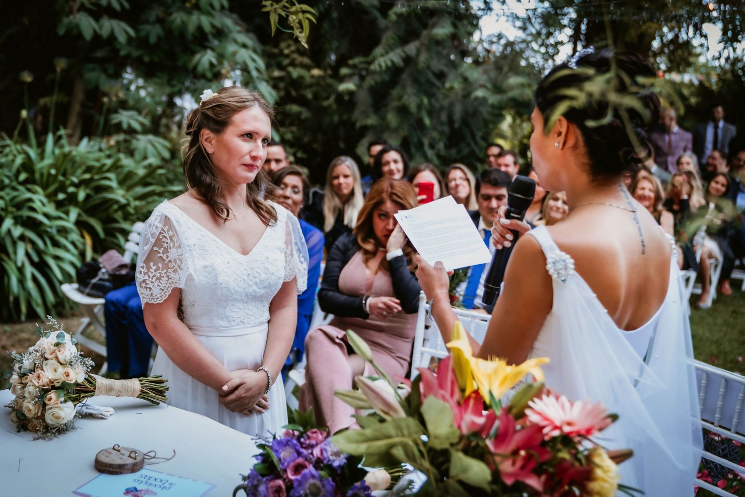 Two brides exchange vows during an outdoor wedding ceremony with guests seated behind.