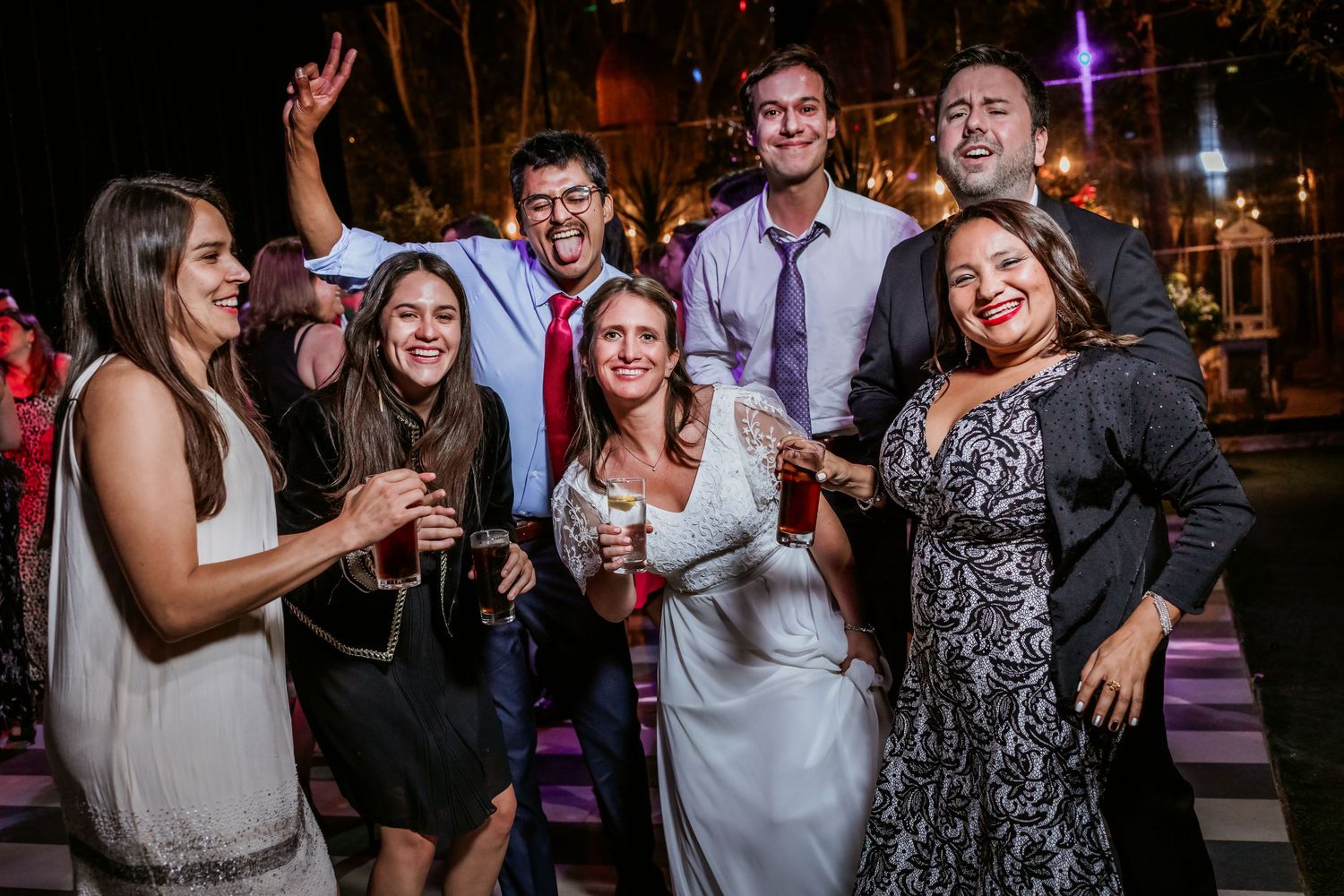 Bride celebrates with group of smiling wedding guests holding drinks at outdoor evening reception.