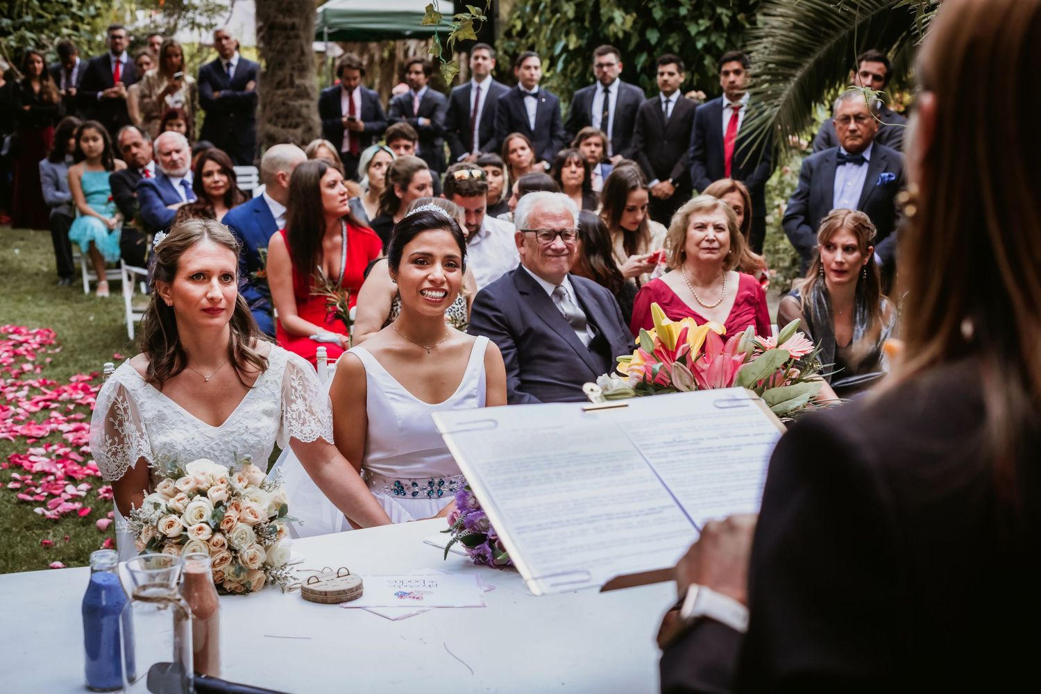Two brides sit at ceremony table with officiant and guests during outdoor garden wedding.