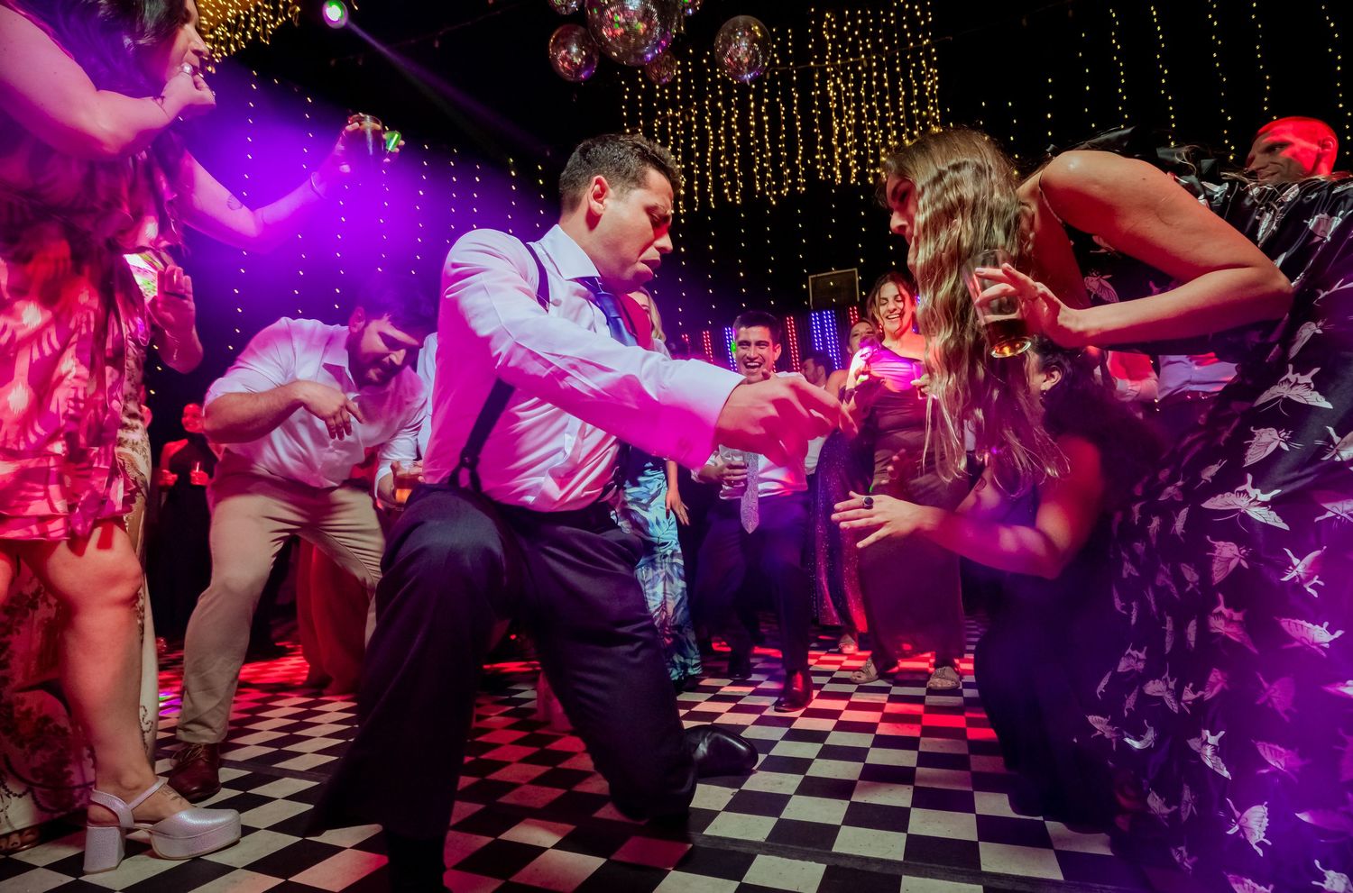Couple dances energetically on a checkered dance floor surrounded by guests at a wedding party.