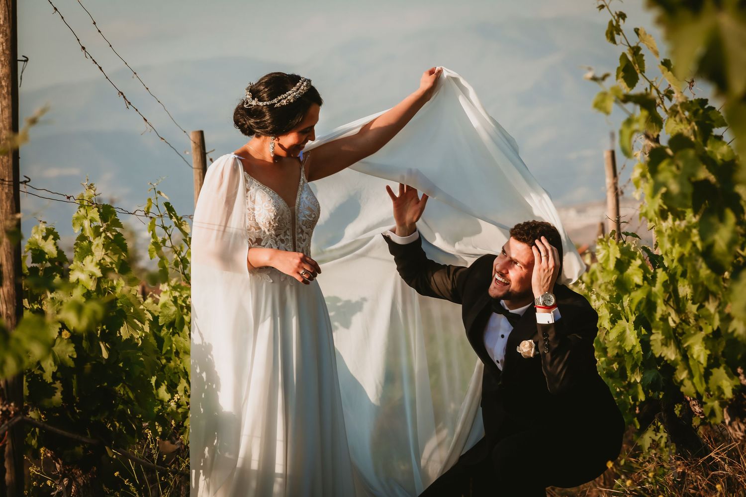 Playful wedding photo of bride lifting her cape veil over groom sitting in a sunny vineyard.