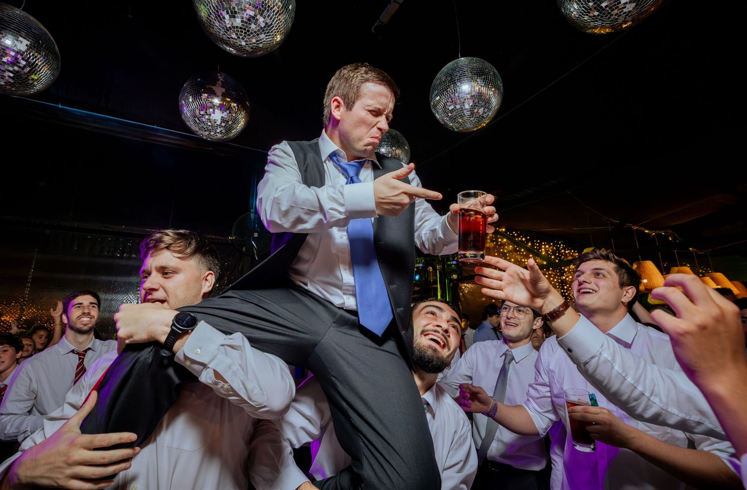 Groomsman lifted on shoulders holding drink surrounded by cheering friends at wedding reception party.