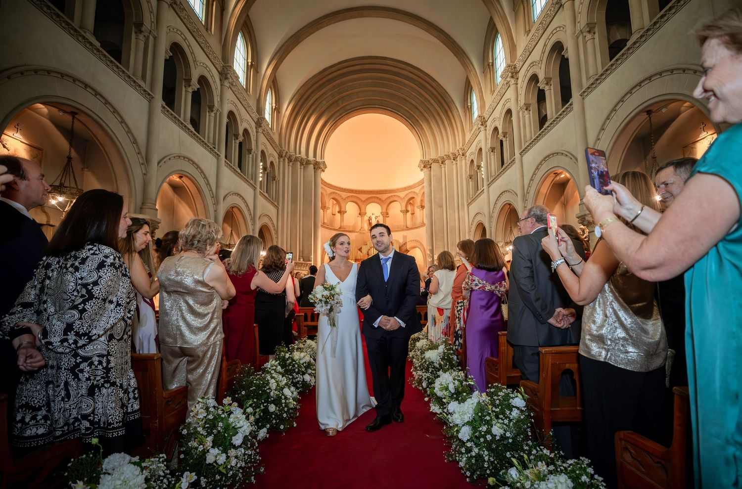 Bride and groom walk down red carpet aisle past cheering guests after church wedding ceremony.
