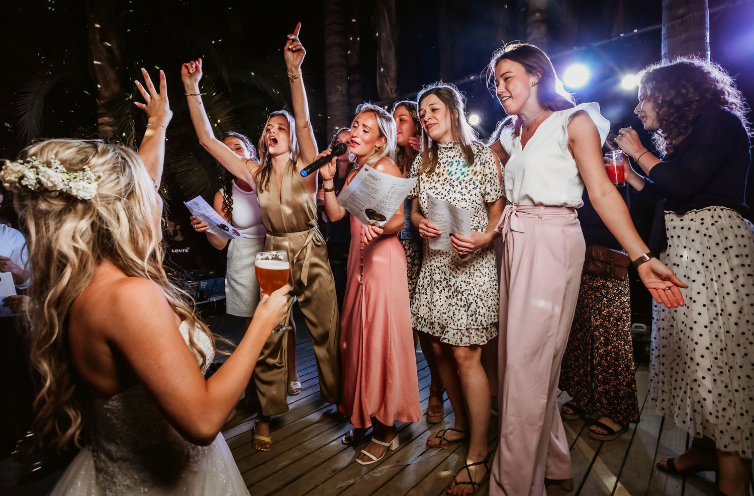 Bride and bridesmaids singing and dancing together at lively outdoor wedding reception at night.