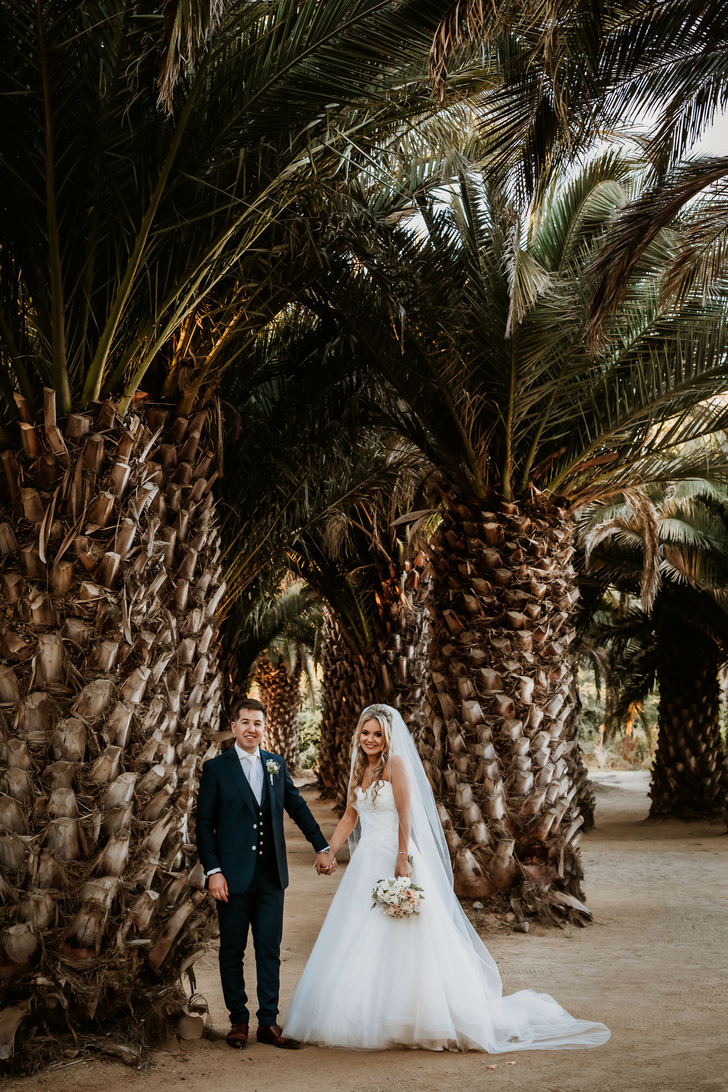 Bride and groom holding hands while walking through a stunning palm tree grove on wedding day.