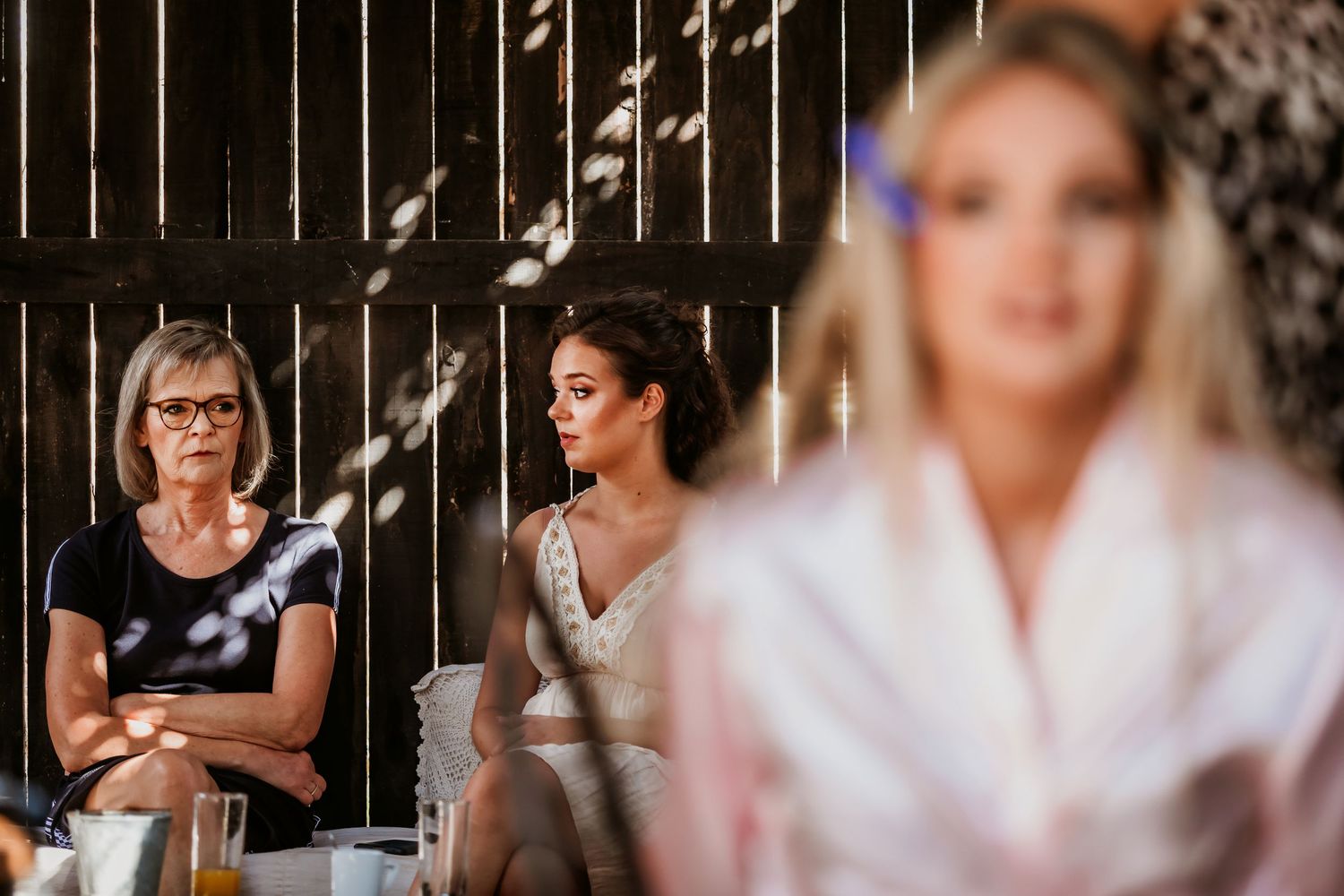 An older woman with glasses sits with arms crossed while two younger women in bridal attire are visible nearby.