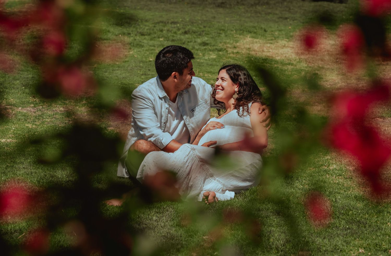 Happy couple sitting on green grass laughing together during outdoor maternity photo session in park.
