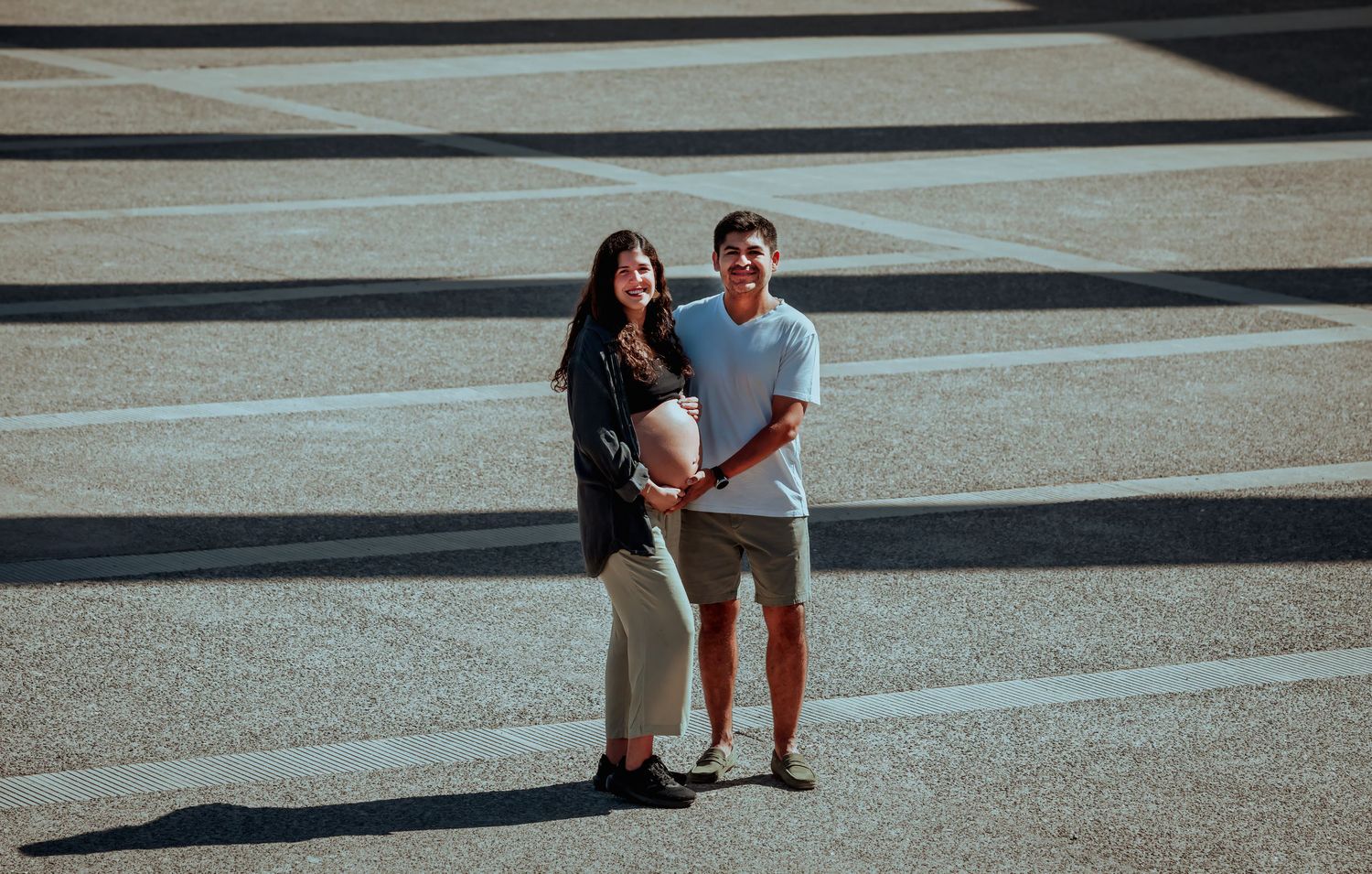 Smiling couple posing together on a sunny striped pavement, woman showing her pregnant belly.