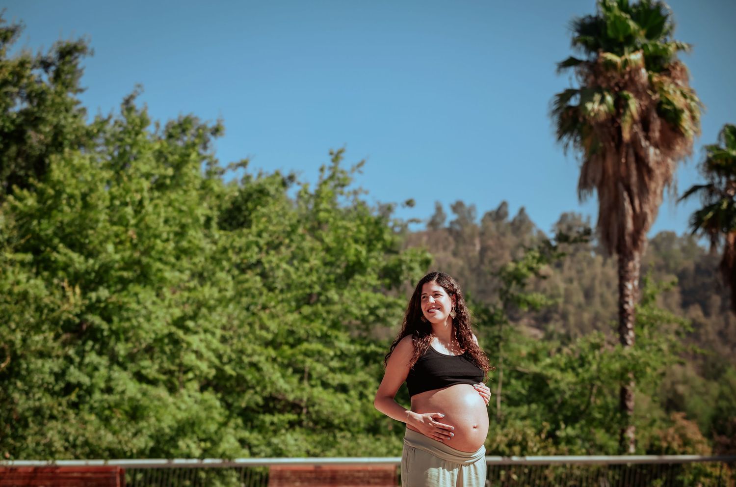 Radiant pregnant woman cradling her bare bump outdoors with palm trees and blue sky behind her.