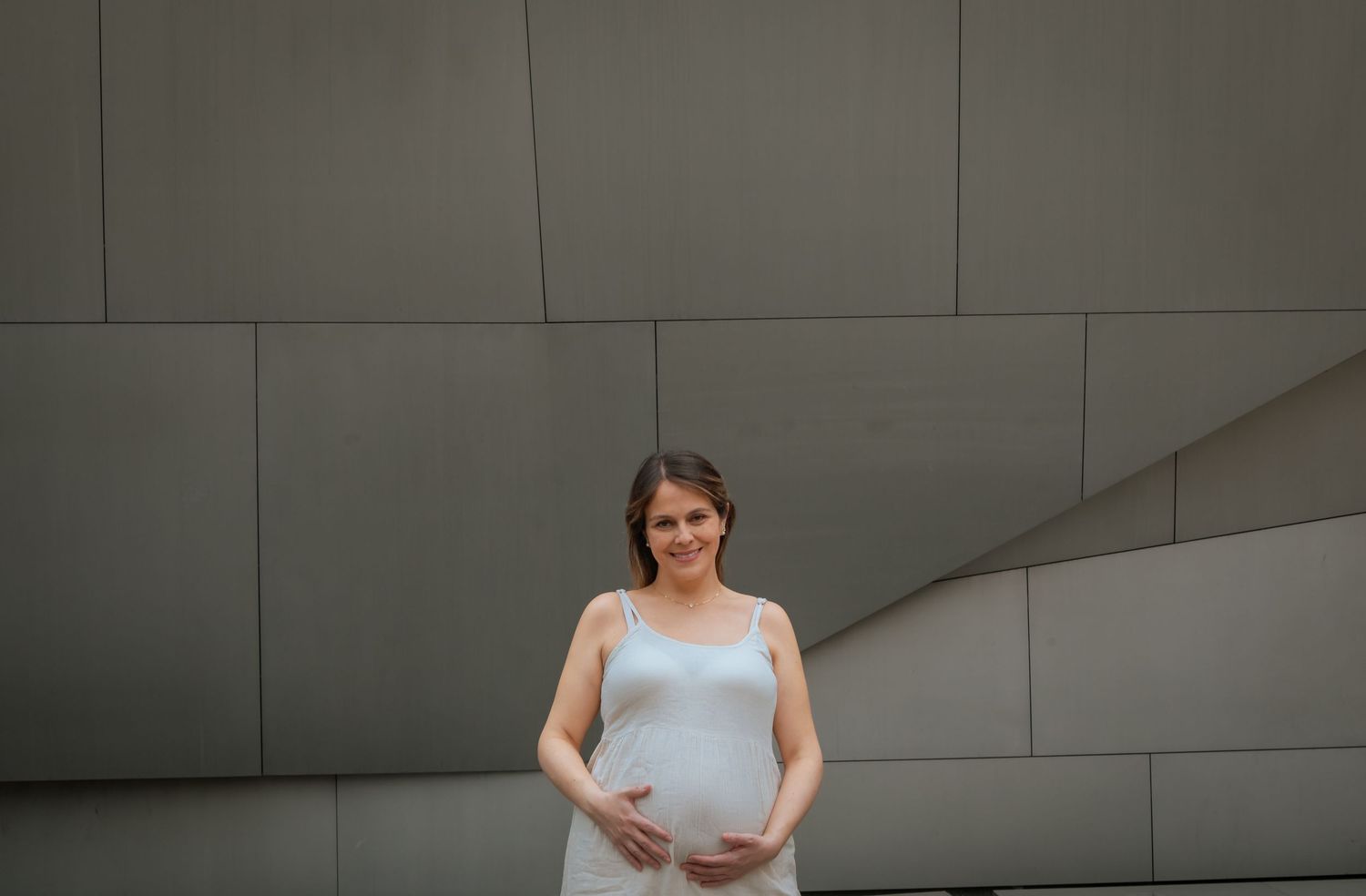 Smiling pregnant woman in a white dress cradling her baby bump while standing against a gray geometric wall.