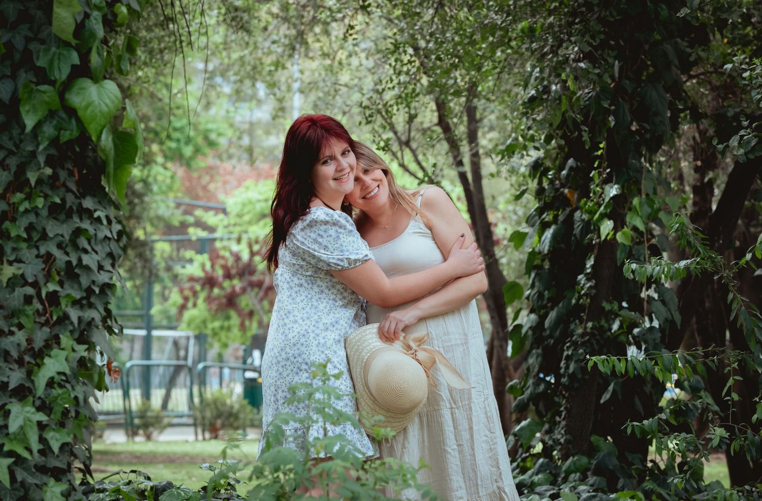Two women share a warm embrace surrounded by lush green garden foliage during an outdoor maternity photo session.