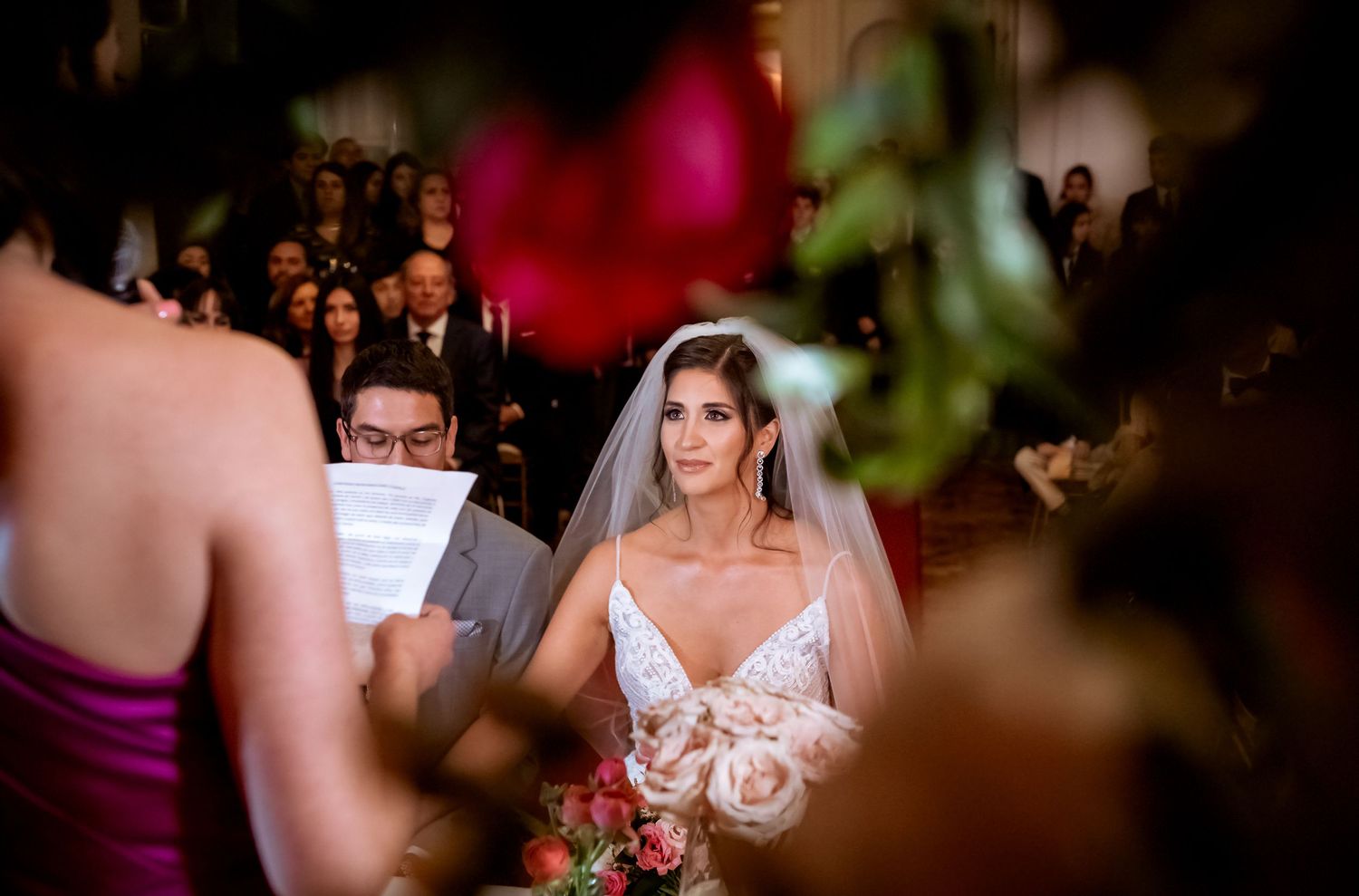Bride in lace wedding gown and veil stands at altar during ceremony, guests seated in elegant venue behind her.