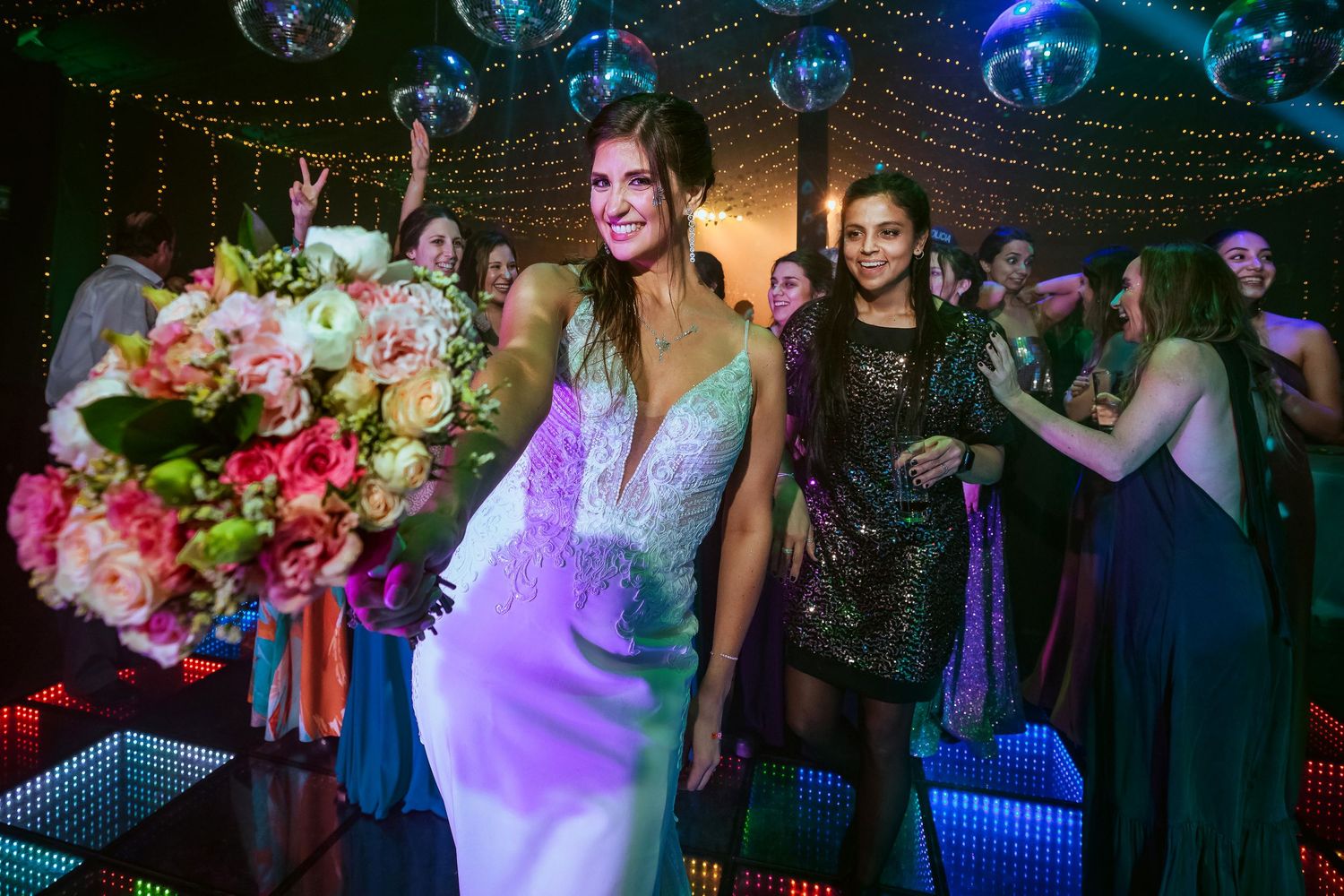 Bride in white gown holds colorful bouquet on dance floor surrounded by guests at a lively wedding reception.