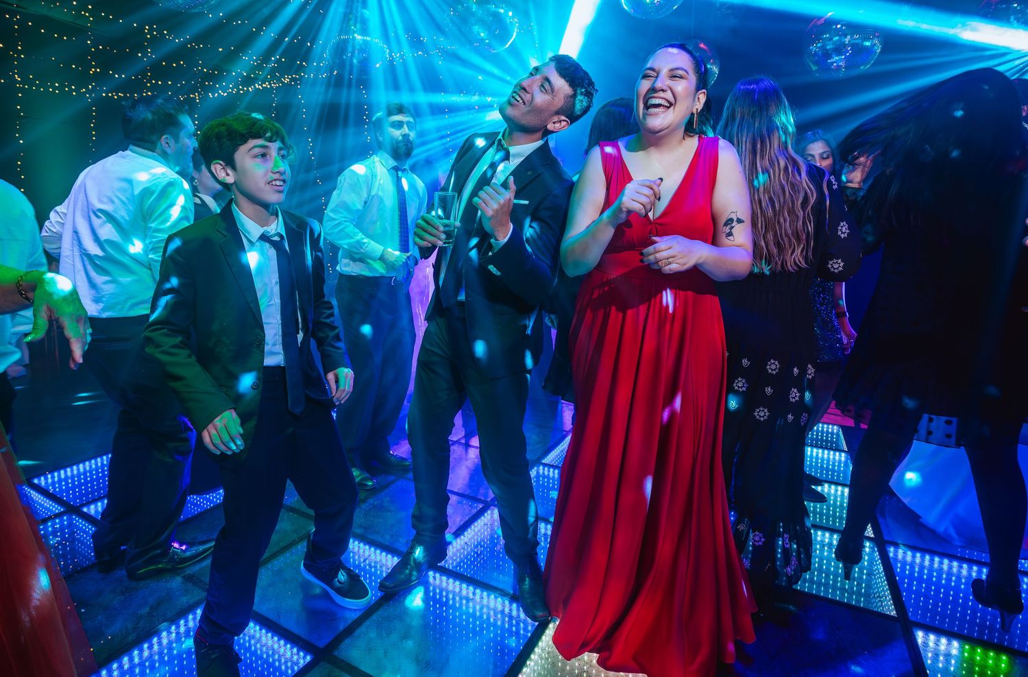 People dancing joyfully at a party, woman in red dress laughing with men in suits on illuminated dance floor.