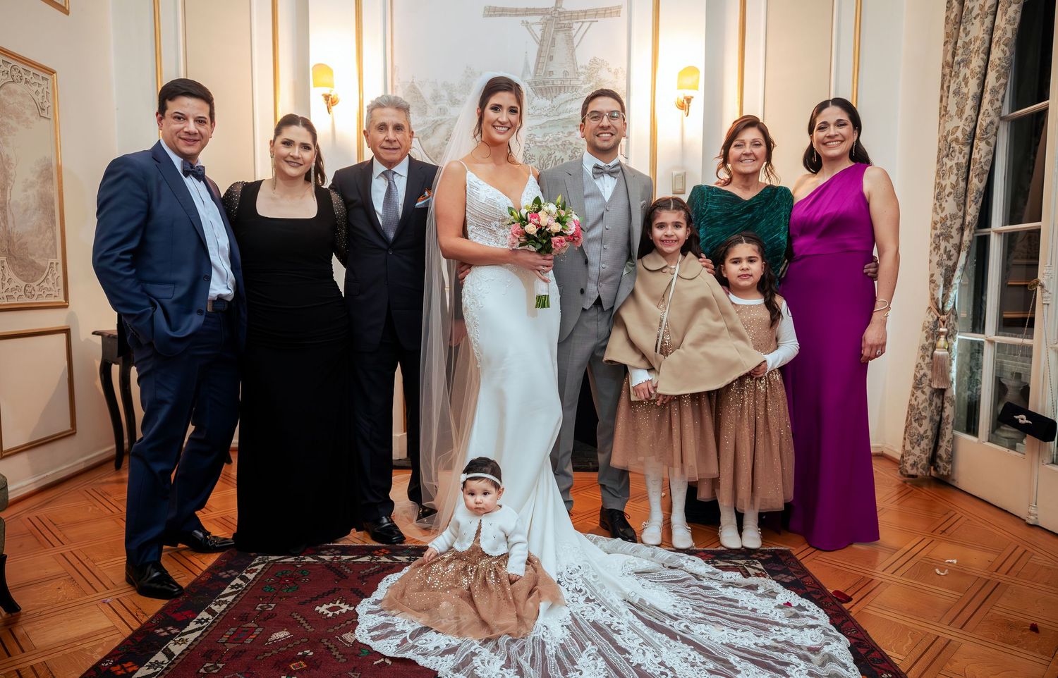 Wedding party poses indoors, bride in white gown with bouquet, groom, family members in formal attire, two flower girls in brown dresses.