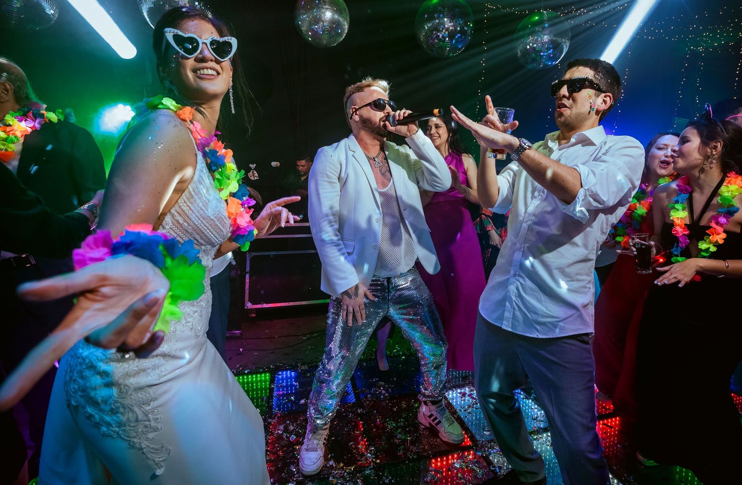 Three performers singing at a vibrant nightclub party with disco lights, lei garlands, and confetti.